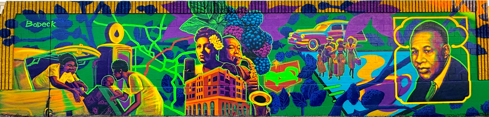 “Safe Travels” by artist Bodeck Luna at the corner of 43rd and Flower Streets. Painted in Luna’s favored hues of green and purple, “Safe Travels” brings focus to Black families traveling in Los Angeles, under the guidance of Victor H. Green’s “Green Book,” a national travel guide first published in 1936 that assisted many Black Americans navigate oppression during the Jim Crow Era. A portrait of Green is seen on the right side of the mural.  The mural also references The Dunbar Hotel, an entry in the “Green Book” as an anchor for traveling musicians and the Black community, with portraits of Billie Holiday and Charlie Parker who frequented the hotel. The depiction of a blackberry branch above Holiday and Parker is a nod to Wallace Thurman’s novel, “The Blacker the Berry” which explores the harsh conditions and prejudices experienced by Black Americans. 