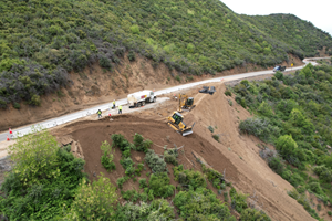 State Route 33 Slope Repair. The image is an aerial view looking down on state route 33 that transverses left to right in the middle of the picture . The  mountains on both sides of the road are green. On the road we see 3 workers on the ground a water truck and a large dump truck.  The slope has fresh material that is being moved by two earth movers.