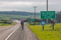 A person riding a bicycle on a road next to a sign. San Simeon