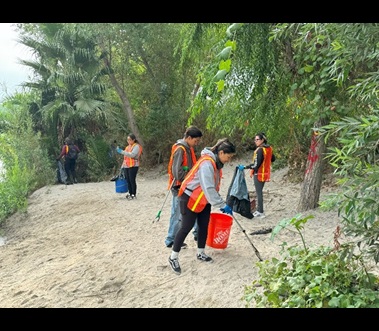 Picture shows a group of six volunteers wearing bright orange vests and large orange paint buckets picking up litter in the sand along a creek bed. They are surrounded by green trees and bushes. 
