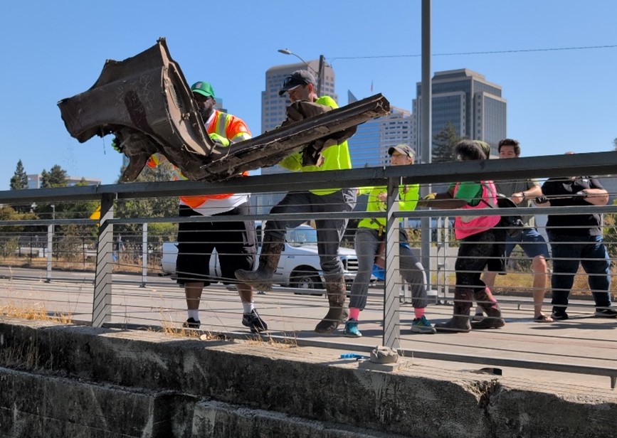 Picture shows a group of volunteers on top of a concrete bridge hoisting up a large, rusted metal piece of a car over the railing of the bridge. Tall buildings and blue sky are in the background. 
