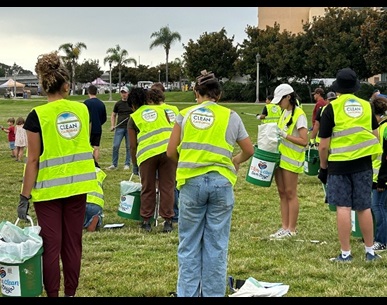 Picture shows a group of about 20 volunteers in bright green vests and large green buckets picking up litter in the grass. In the background are trees and a park. 