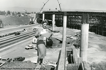 Black and white photo of a freeway construction site with a woman dressed in 1950s era skirt suit looking at plans in foreground.