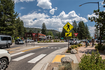 Photo of “complete street” transportation infrastructure with vehicles navigating a roundabout. A car has stopped at a crosswalk that has pedestrian and ”Yield” signage and a man and boy ride their bikes in an adjacent protected bike/pedestrian path. There is a silver; metal sculpture in the middle of the roundabout and the project’s landscaping incorporates granite rocks and low-water plantings. Views of a blue sky; businesses; pine trees and the Sierra Nevada mountains complete the background.