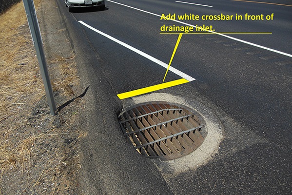 From above, a view of a road surface with a bike lane bending to circumnavigate a sewer drain.
