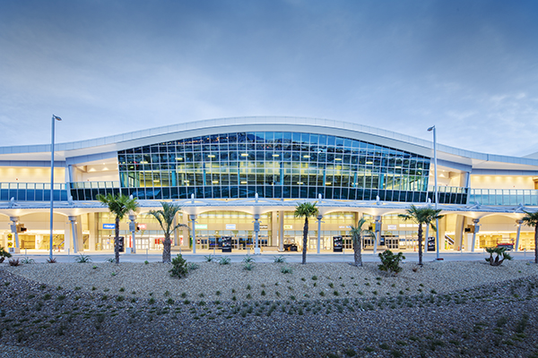 A view of San Diego International Airport Rental Car Center. The building looks well-lit and has a rounded curving roof angling over the main entrance with palm trees interspersed in the foreground. 