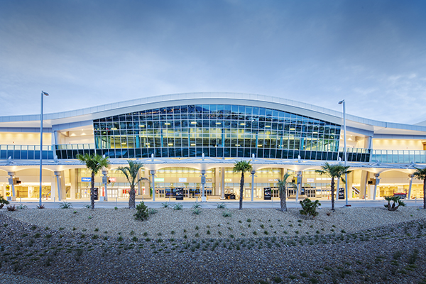 A view of San Diego International Airport Rental Car Center. The building looks well-lit and has a rounded curving roof angling over the main entrance with palm trees interspersed in the foreground.