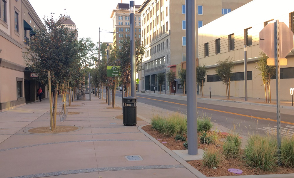 An city street with trees planted at even intervals on a sidewalk with a unique paving and brick pattern.