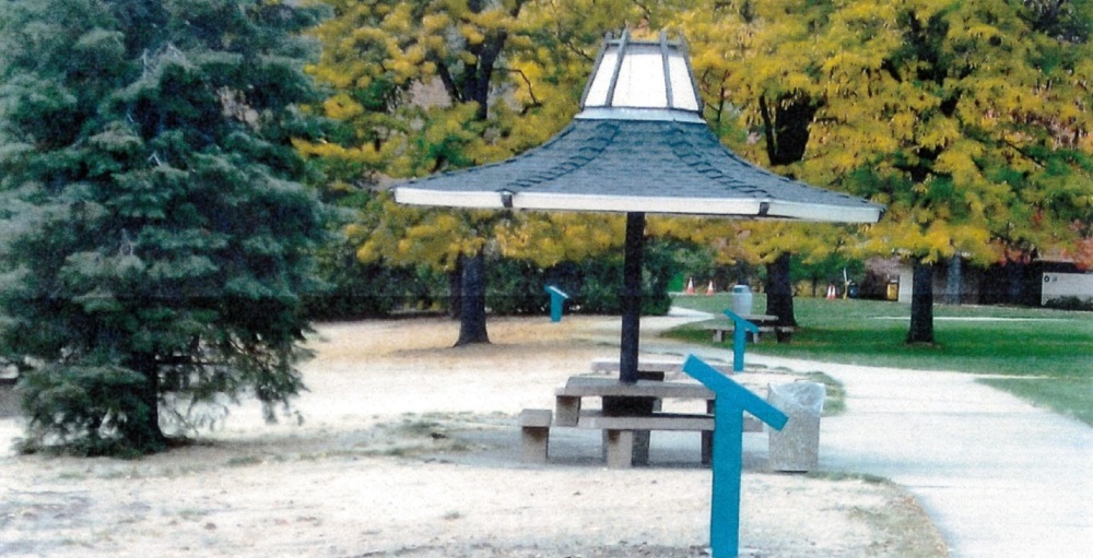 A side-view of a rest area with signage and a gazebo with picnic table underneath it. Trees are in the background.