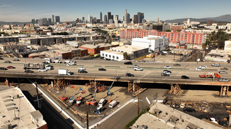 An aerial photo taken on Nov 21, 2023, from above a section of Interstate 10 at Alameda Street in Downtown Los Angeles where there was a fire on Nov 11; burn marks are still visible on the ground. The image shows the ongoing repair work happening while the freeway is open in both directions. Workers on a piece of equipment with a raised platform work on a guardrail from the outside edge of the structure.