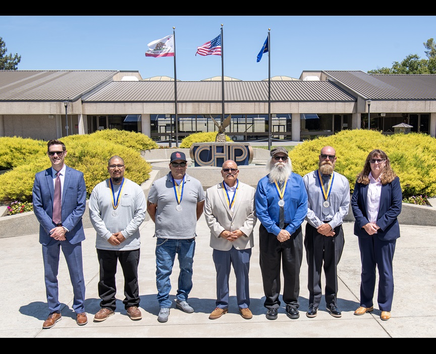 Picture shows six men and one woman standing in a line in front of the California Highway Patrol Academy in West Sacramento. Behind them is the CHP Logo with the California and American flags on flag poles. The people in the photo are  CalSTA Undersecretary James Hacker Isai Camino John Bedolla Jeronimo Prado Vernon Ladd Jarett Walter Lopez Janice Benton  