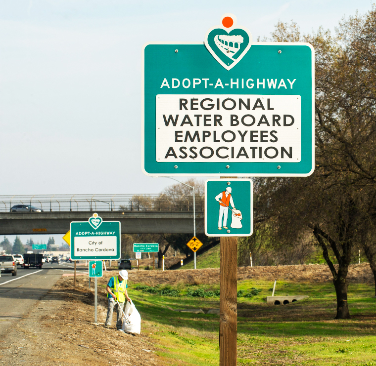 Photo of an Adopt-A-Highway sign along right of way. Freeway overpass and a person cleaning up litter along the roadway are visible in the background.