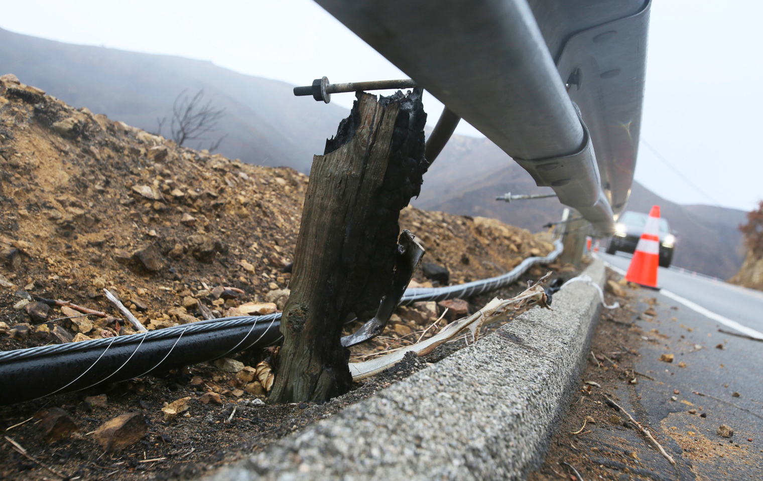 Photo of burned vegetation and damaged guard rail along a highway. A car and safety cone in the background.