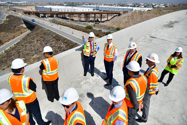 Photo of many people wearing Caltrans hard hats, standing on an overpass at the Otay Mesa East construction site.