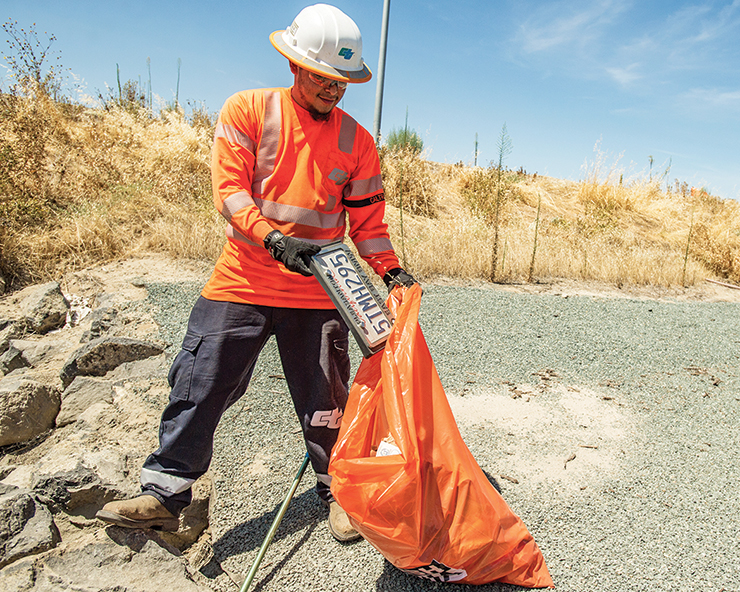 Photo shows a Caltrans maintenance worker disposing of trash into a plastic bag along the State highway