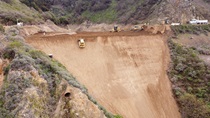 California Highway 1 at Rat Creek near Big Sur: aerial view of large construction equipment dwarfed by the enormity of the work site. One piece of equipment is being deployed in a precarious-looking vertical position on the ocean side of the coast just below the washed out road level. 