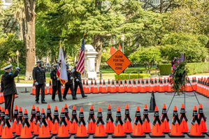 Caltrans honor guard members saluting US and California flags during the 2021 Caltrans Workers Memorial ceremony in Sacramento, California's Capitol Park to honor fallen highway workers. They are surrounded by orange cones wrapped in black bands with the names of each of the 189 fallen workers. The cones are arranged in the shape of a diamond, mimicking the orange "Slow for the Cone Zone" sign in the middle. 