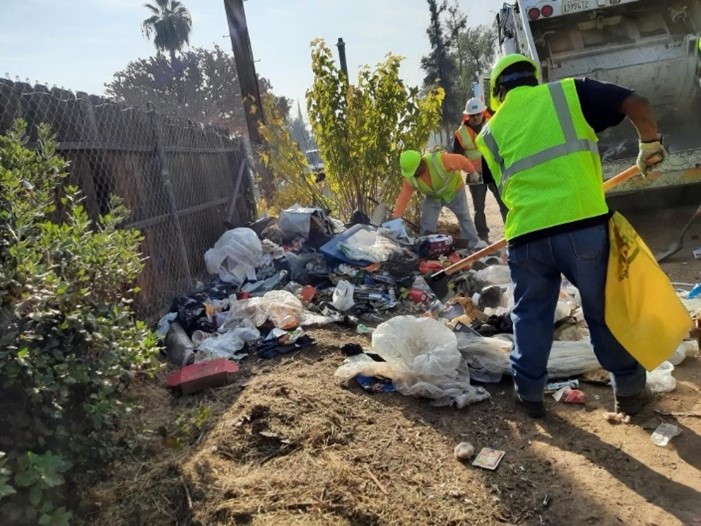 Three workers in fluorescent yellow vests and white or yellow hardhats add trash to a pile between a fenceline and the garbage truck that will haul it away.