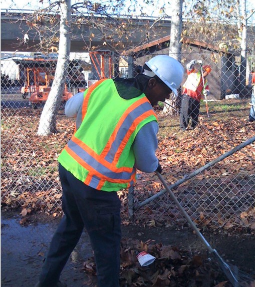 Near a chain link fence, two workers in reflective yellow vests and white hardhats rake up leaves and litter for removal. 
