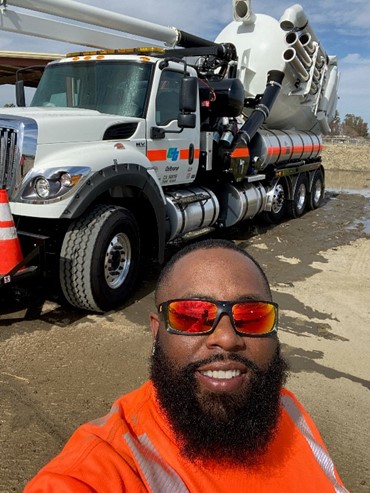 Maintenance Worker Robert Kelso smiles in a reflective fluorescent orange sweatshirt and sunglasses as he poses in front of a large Caltrans truck.