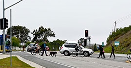 Walkers and bikers share the road with vehicles in Caltrans District 5, which covers California’s Cental Coast. They cross the street at a crosswalk as a white SUV drives in the same direction in the background.