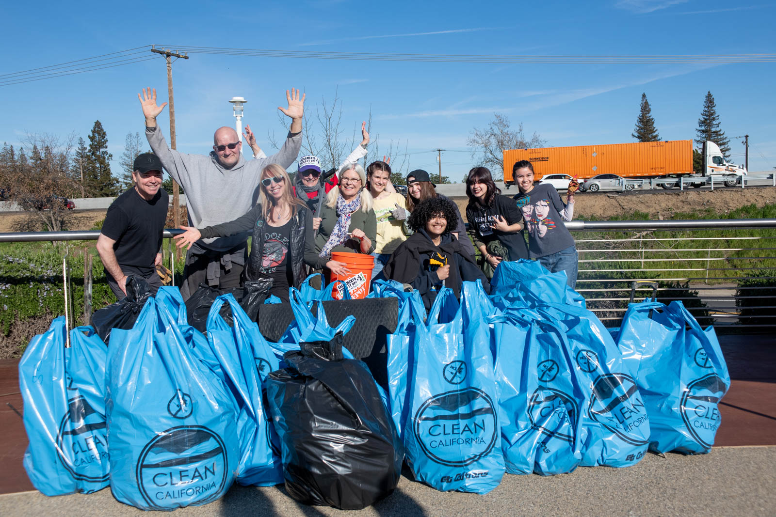 Volunteers from the Sacramento Kings NBA organization and Sacramento Picks It Up! join the Caltrans Stormwater and Clean California campaign teams for a celebratory group photo behind a pile of more than a dozen large, blue, Clean California-branded garbage bags filled with the trash they picked up at the Clean California Sacramento River Clean Up event. 