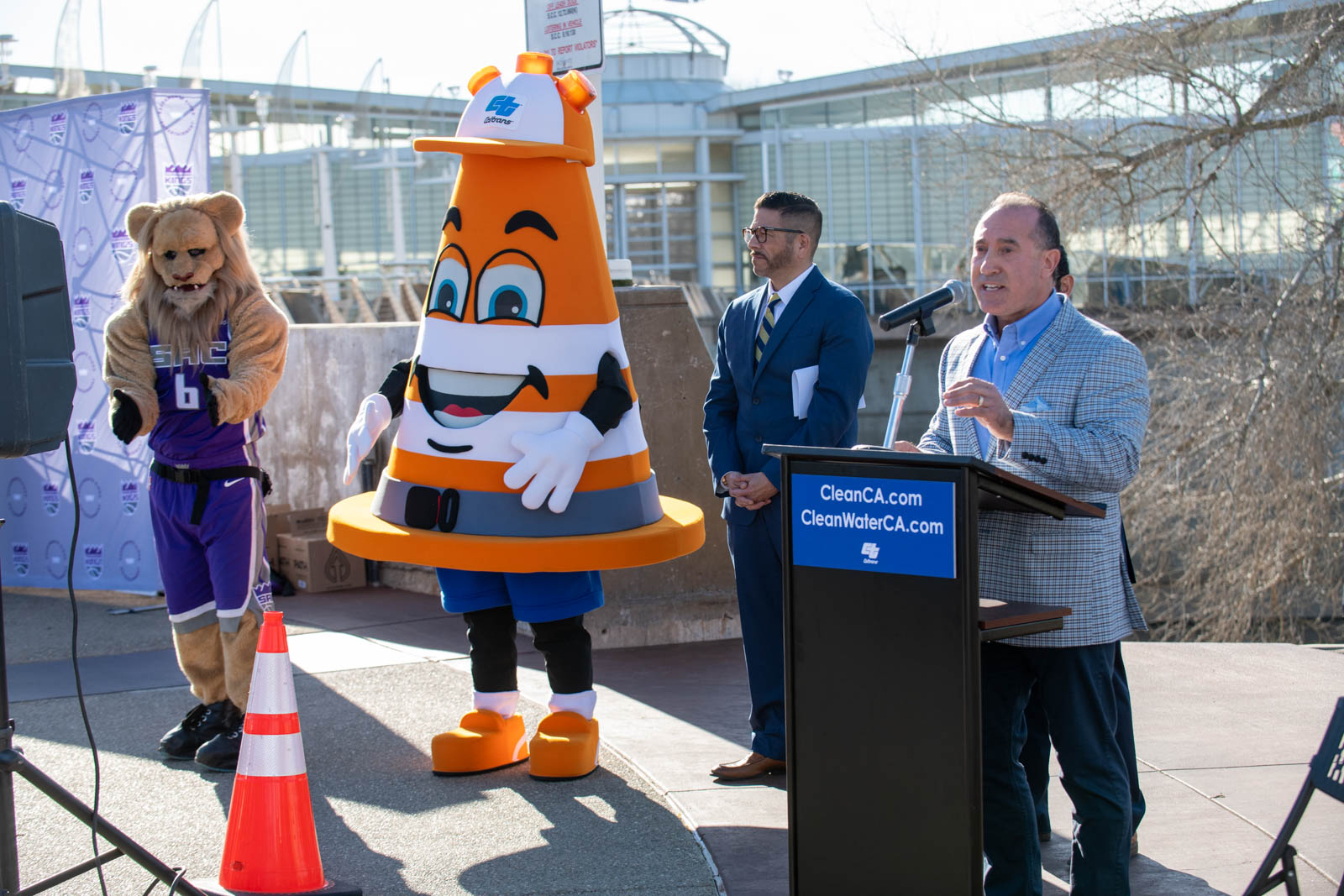 Caltrans Director Tony Tavares addressed the crowd at the Clean California Sacramento River Clean Up event from behind a dark brown wooden podium. The blue sign on the podium reads “CleanCA.com” and “CleanWaterCA.com” with the Caltrans logo below, in white. Standing to the Director’s right: Sampson the lion mascot, wearing a purple NBA Sacramento Kings team uniform with the number 6; Safety Sam, Caltrans’ orange cone mascot wearing a hardhat; Loren Magaña, Caltrans’ Stormwater campaign manager; and obscured by the Director, Sacramento Vice Mayor Eric Guerra.