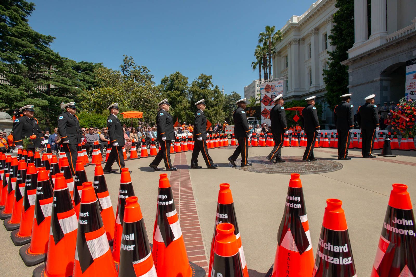 Photo of the 2022 Caltrans Workers Memorial Ceremony. Orange cones with black name bands representing the 189 fallen state highway workers who died in the line of duty since 1921 are arranged in a diamond shape like a caution sign. Inside the diamond, uniformed Caltrans Honor Guards place a ceremonial black cone in front of a floral wreath during the memorial.