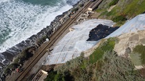 Color aerial photo down a steep San Clemente hillside where a landslide halted passenger train service between the California cities of Irvine and Oceanside on Jan 24, 2024. Workers have installed construction materials over the affected area to keep more debris from tumbling onto the railroad tracks below. At the bottom of the photo, workers and a vehicle are visible by the tracks that run parallel to the Pacific Ocean.