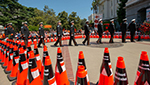 Photo of the 2022 Caltrans Workers Memorial Ceremony. Orange cones with black name bands representing the 189 fallen state highway workers who died in the line of duty since 1921 are arranged in a diamond shape like a caution sign. Inside the diamond; uniformed Caltrans Honor Guards place a ceremonial black cone in front of a floral wreath during the memorial.